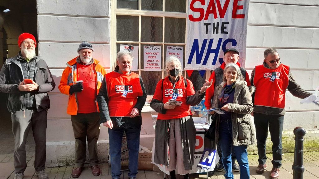A group of seven campaigners in red T-shirts stands on a sunny street with a large placard behind them reading Save the NHS