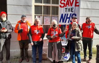 A group of seven campaigners in red SOHS T-shirts stands on a sunny street with a large placard behind them reading Save the NHS